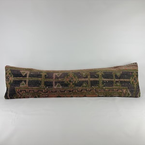 May include: A long, rectangular lumbar pillow featuring a dark, weathered textile cover with intricate geometric patterns in shades of deep purple, muted green, and faded orange, finished with a textured, woven appearance.
