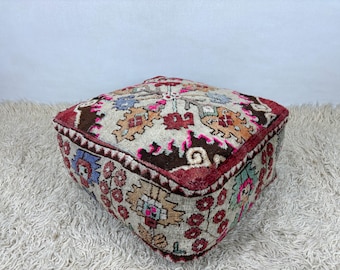 May include: A square, patterned pouf with a beige background and a colorful floral design. The edges are a deep burgundy, and the top features a complex pattern with pink, orange, and blue accents. The pouf is placed on a shaggy, white rug.