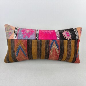 May include: A rectangular throw pillow featuring a top band of multicolored geometric patterns in pink, purple, and black, and a bottom section with vertical brown, tan, and red stripes accented by blue and beige chevron-style embroidered motifs.