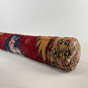 May include: A cylindrical bolster pillow featuring a woven textile cover with a deep red base colour, accented by patches of dark blue, mustard yellow, and tan. The fabric displays a textured, knotted pile surface with intricate geometric patterns.