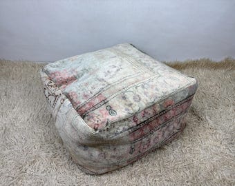 May include: A square floor pouf featuring a faded, distressed textile surface with a muted color palette of cream, soft pink, and grey. The fabric displays intricate, worn-in patterns and geometric borders, creating a textured, low-profile seat.