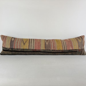 May include: A long, rectangular pillow with a colorful, striped design. The pillow features a mix of colors including pink, yellow, brown, and black. The pillow is likely made of a woven fabric and is a decorative item.