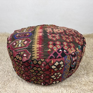 Puede incluir: Un puf de suelo redondo con una superficie textil tejida con un patrón geométrico multicolor. El diseño incorpora tonos de rojo intenso, marrón oscuro, negro, azul y crema, dispuestos en formas repetitivas y bandas verticales.