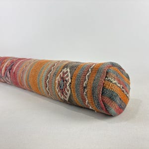 Può includere: Un cuscino cilindrico con una fodera in tessuto intrecciato caratterizzato da un motivo a righe multicolore nei toni dell'arancione, grigio, rosso e crema, arricchito da un motivo floreale geometrico ripetuto lungo la superficie del tessuto.
