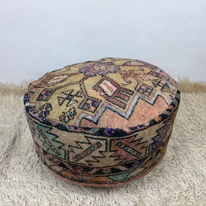 May include: A round floor pouf featuring a woven textile surface with a multi-colored geometric pattern in shades of tan, peach, black, green, and purple. The cylindrical ottoman has a flat top and vertical sides with a dense, textured weave.