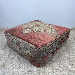 Pode incluir: Um pufe de chão quadrado com uma capa de tecido com um padrão geométrico vermelho e creme desbotado na parte superior e um motivo repetitivo cinzento, vermelho e creme nas laterais, construído com uma forma baixa, quadrada e firme.
