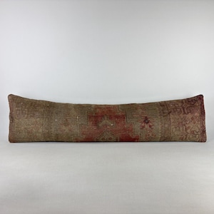 May include: A long, rectangular bolster pillow featuring a faded, distressed-look textile cover. The fabric displays a complex, symmetrical pattern in muted shades of rust red, beige, and brown, with a worn, textured appearance across the surface.