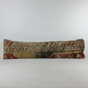 May include: A long, rectangular bolster pillow featuring a multi-colored woven pattern in shades of red, beige, brown, and black. The textile displays intricate geometric and floral motifs across its entire surface, with a textured appearance.