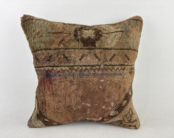 May include: Square decorative pillow with a distressed, vintage look. The pillow features a brown and tan color palette with geometric and floral patterns. The fabric has a textured appearance, suggesting a woven or rug-like material.