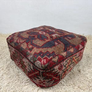 Peut inclure: Pouf carré à motifs dans les tons de rouge, marron et bleu. Le textile présente un motif géométrique avec des éléments floraux. Le pouf est posé sur un tapis blanc et duveteux.