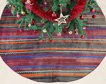 Pode incluir: Uma saia de árvore de Natal redonda com um padrão listrado em tons de vermelho, laranja, azul e cinza. A saia tem uma borda branca em zigue-zague. A árvore é decorada com enfeites vermelhos e dourados, uma guirlanda vermelha e uma estrela de madeira.