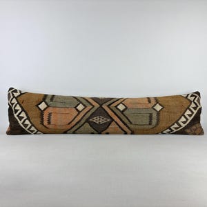 May include: A long, rectangular lumbar cushion featuring a woven textile cover with a geometric pattern in shades of brown, tan, muted green, and cream. The design includes diamond shapes and angular lines, finished with a dark brown border.