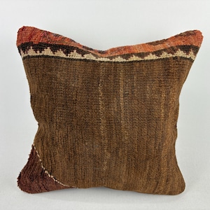 May include: A square, textured pillow with a brown body and a decorative top border. The top border features a geometric pattern in shades of orange, red, and cream. The pillow has a rustic, handcrafted appearance, suitable for home decor.