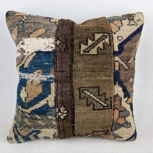 May include: A square throw cushion featuring a patchwork design of woven textile fragments. The surface displays a mix of blue, brown, and beige tones with geometric patterns, including a central vertical brown panel with dark zigzag motifs.