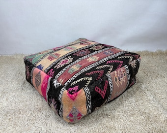May include: A square floor cushion featuring a woven textile cover with a multi-colored geometric pattern. The design includes alternating stripes of black, cream, pink, and green, decorated with intricate diamond and chevron motifs throughout.
