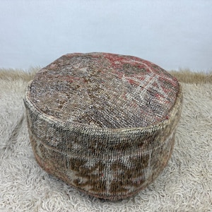 Può includere: Un pouf da pavimento rotondo realizzato in tessuto vintage intrecciato con un motivo usurato nei toni del marrone, beige e rosso sbiadito. Il pouf cilindrico presenta una superficie superiore piatta e lati verticali con finitura annodata.