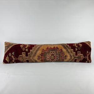 May include: A long, rectangular bolster pillow featuring a deep burgundy and tan woven pattern. The design displays intricate geometric shapes and floral motifs centered on the front, with a textured fabric finish throughout the entire surface.