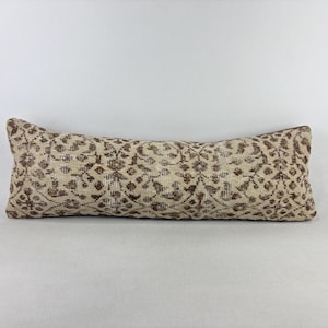 May include: A long, rectangular lumbar pillow featuring a repeating brown floral and geometric pattern set against a light beige, textured fabric background. The pillow has a structured, elongated shape with visible woven details across the surface.