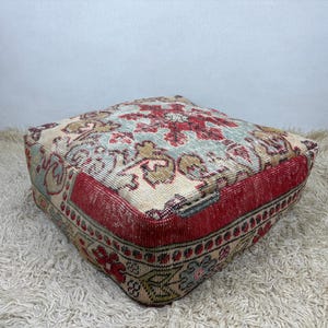 May include: A square floor pouf featuring a woven textile cover with a complex geometric and floral pattern in shades of red, cream, and light blue. The sides are finished with a solid red border and a repeating decorative motif along the base.