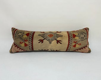 May include: A long, rectangular pillow with a beige background and a colorful geometric pattern. The pattern features red, blue, and green colors and includes a central diamond shape.