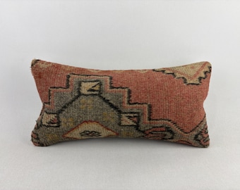 May include: Rectangular decorative pillow with a geometric pattern in shades of red, gray, and beige. The pillow has a textured appearance, suggesting a woven or embroidered design. The pattern features angular shapes and lines.