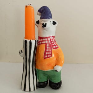 Könnte beinhalten: Ein skurriler Kerzenhalter aus Keramik mit einem Eisbären-Design. Der Bär trägt eine blaue Mütze, ein orangefarbenes Hemd, einen rot-weiß karierten Schal und grüne Hosen. Der Kerzenhalter ist schwarz-weiß gestreift und hält eine orangefarbene Kerze.
