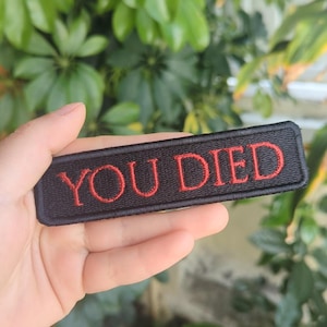 May include: Black rectangular patch with the embroidered text "YOU DIED" in red. The patch is held in front of a background of green foliage.