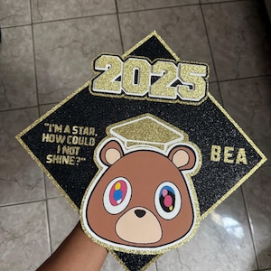 May include: Black graduation cap with gold glitter accents. Features a brown bear graphic with colorful eyes and the text "2025". The quote "I'm a star, how could I not shine?" is also included.