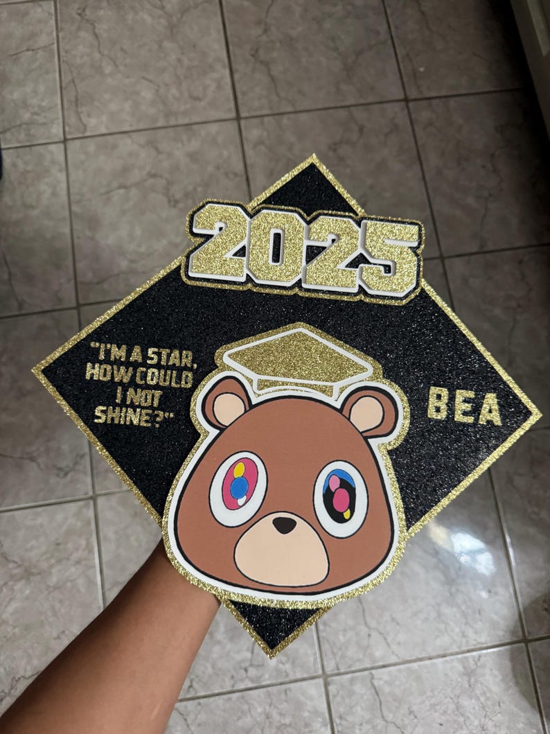 May include: Black graduation cap with gold glitter accents. Features a brown bear graphic with colorful eyes and the text "2025". The quote "I'm a star, how could I not shine?" is also included.