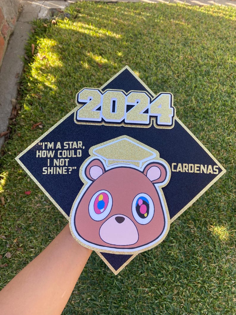 May include: Black graduation cap with gold glitter trim and a cartoon bear graphic. The cap features the year "2024" in gold glitter and the name "Cardenas". A quote that reads "I'M A STAR, HOW COULD I NOT SHINE?" is also visible.
