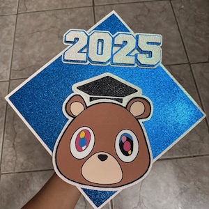 May include: A graduation cap with a blue glitter diamond shape and the year "2025" in silver glitter. A brown bear face with colorful eyes and a graduation cap is centered on the cap.