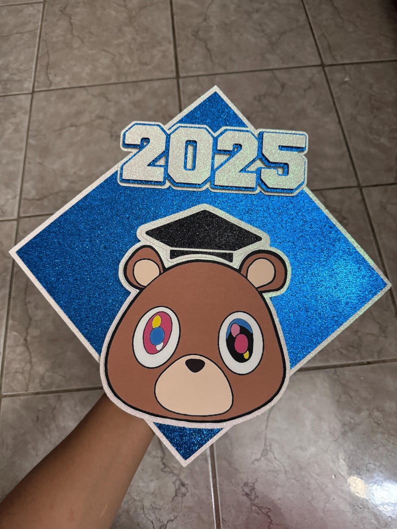May include: A graduation cap with a blue glitter diamond shape and the year "2025" in silver glitter. A brown bear face with colorful eyes and a graduation cap is centered on the cap.
