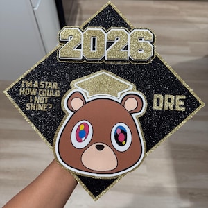 May include: Black graduation cap with gold glitter accents and the year "2026". Features a brown bear graphic with colorful eyes and the text "I'M A STAR, HOW COULD I NOT SHINE?" and "DRE".