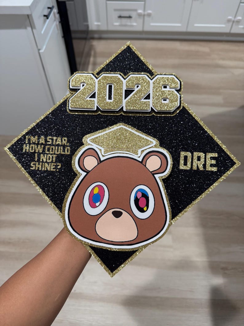 May include: Black graduation cap with gold glitter accents and the year "2026". Features a brown bear graphic with colorful eyes and the text "I'M A STAR, HOW COULD I NOT SHINE?" and "DRE".