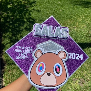 May include: A purple glitter graduation cap with the name "SALAS" in silver letters. The cap features a cartoon bear with colorful eyes and the quote "I'M A STAR, HOW COULD I NOT SHINE?" and the year 2024.