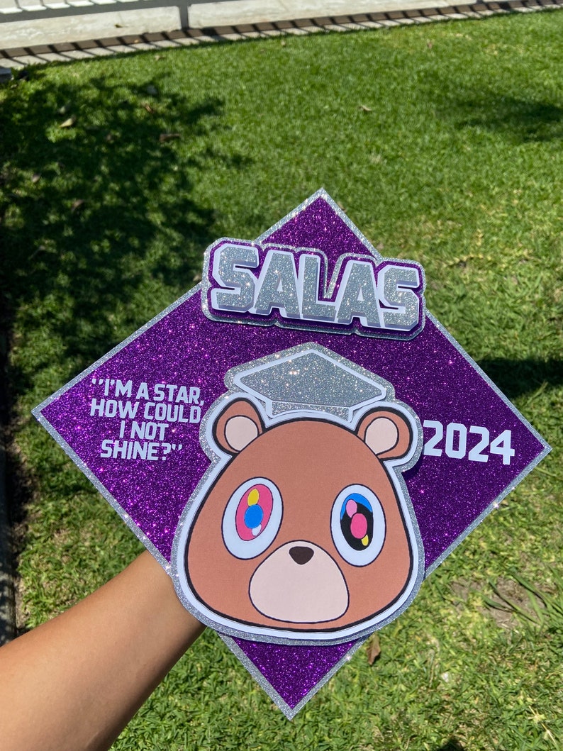 May include: A purple glitter graduation cap with the name "SALAS" in silver letters. The cap features a cartoon bear with colorful eyes and the quote "I'M A STAR, HOW COULD I NOT SHINE?" and the year 2024.