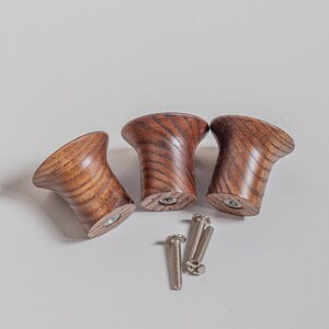 May include: Three wooden cabinet knobs with a rich, warm wood grain pattern. The knobs have a flared, conical shape and are accompanied by several silver screws, suggesting they are ready for installation. These knobs are a decorative hardware element.