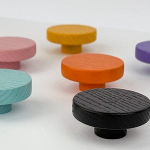 Può includere: Una collezione di otto pomelli in legno colorati in varie tonalità, tra cui viola, rosa, giallo, arancione, verde acqua, nero e blu navy. Ogni pomello ha una parte superiore rotonda con un motivo a grana strutturata e una base cilindrica corta.