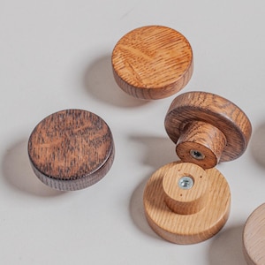 Könnte beinhalten: Sechs Holzschrankknöpfe in verschiedenen Braun- und Beigetönen. Die Knöpfe sind rund mit flacher Oberseite und zylindrischem Sockel, die die natürliche Holzmaserung zeigen. Ein Knopf ist mit einer Schraube an einer Basis befestigt.