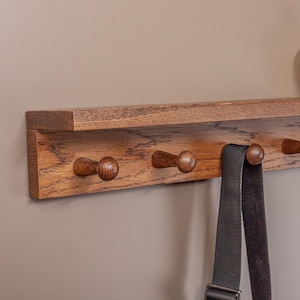 Puede incluir: Estante de pared de madera con seis ganchos y un pequeño estante superior. El estante tiene un acabado de madera natural y sostiene tres jarrones decorativos de color crema. Una correa negra cuelga de uno de los ganchos.