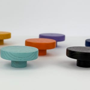 May include: A collection of colorful wooden knobs in various shades, including purple, pink, yellow, teal, orange, black, and blue. Each knob has a round top and a short, cylindrical base. The knobs are arranged on a white surface.