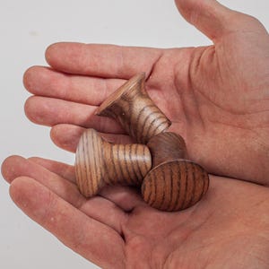 May include: Four wooden cabinet knobs in various shapes, held in open hands against a white background. The knobs are light brown with visible wood grain, and include a mushroom-shaped knob and an oval knob.