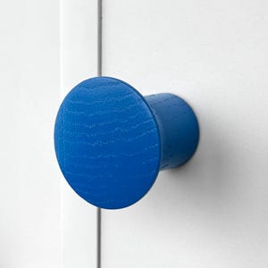 May include: A bright blue wooden cabinet knob with a circular design. The knob is mounted on a white surface, offering a pop of color against the neutral background. The knob's dimensions are approximately 2 inches in diameter.