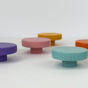 Może przedstawiać: Kolekcja kolorowych drewnianych gałek w różnych odcieniach, w tym fioletowym, różowym, żółtym, turkusowym, pomarańczowym, czarnym i niebieskim. Każda gałka ma okrągłą górę i krótką, cylindryczną podstawę. Gałki są ułożone na białej powierzchni.