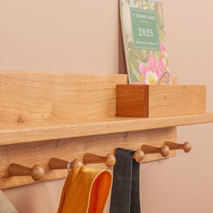 Puede incluir: Estante de pared de madera con una repisa y seis ganchos. Un pequeño cactus en una maceta gris se encuentra en el estante, junto con un calendario. Una bufanda amarilla y una correa negra cuelgan de los ganchos.