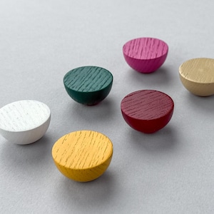Könnte beinhalten: Sechs farbige, halbkugelförmige Holzknebel in Weiß, Grün, Rosa, Rot, Gelb und Beige. Jeder Knebel hat eine strukturierte Holzmaserung. Die Knöpfe sind auf einem hellgrauen Hintergrund angeordnet.