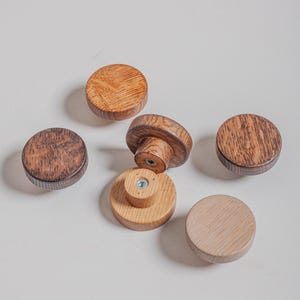 Puede incluir: Un conjunto de seis pomos de madera para armarios en varios tonos de marrón y beige. Los pomos son redondos con una parte superior plana y una base cilíndrica, mostrando la veta natural de la madera. Un pomo está fijado a una base con un tornillo.