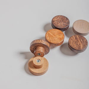May include: A selection of wooden cabinet knobs in shades of brown, from light oak to dark walnut. Some knobs are flat, others have a cylindrical base. The knobs are arranged on a white surface.