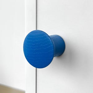 Puede incluir: Un pomo de armario de madera azul vibrante, con una cara circular y una base cilíndrica. El pomo está montado sobre una superficie blanca, mostrando su color brillante y su diseño sencillo. Adecuado para cajones y armarios.