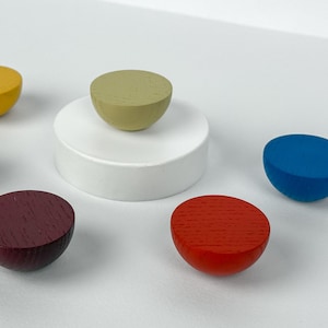 Könnte beinhalten: Sechs halbkugelförmige Holzobjekte in verschiedenen Farben: Gelb, Rosa, Grün, Blau, Rot und Burgunder. Ein grünes Objekt ruht auf einem weißen Sockel. Die Objekte sind auf einer weißen Oberfläche angeordnet.